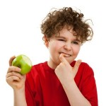 Child holding a green apple and grimacing while trying to remove food stuck in teeth, illustrating common dental hygiene issues.