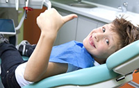 Child in dental chair giving a thumbs-up, wearing a blue bib, in a modern dental office, reflecting positive experience at Water Tower Dental Care.